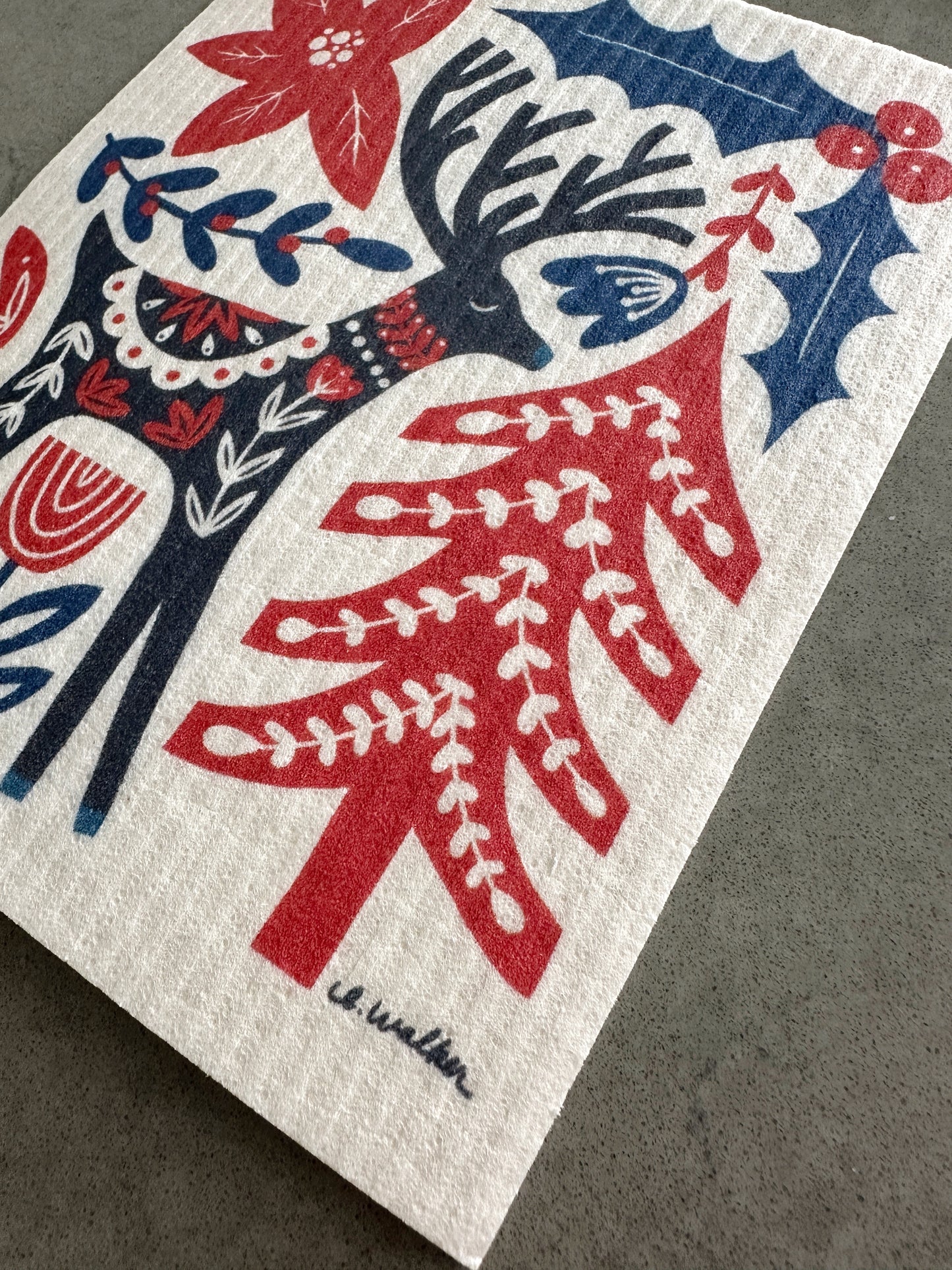 A close-up of a dishcloth featuring a folk-art style design with a red Christmas tree, a dark blue reindeer, and floral motifs on an off-white background. A signature reading e.walker.