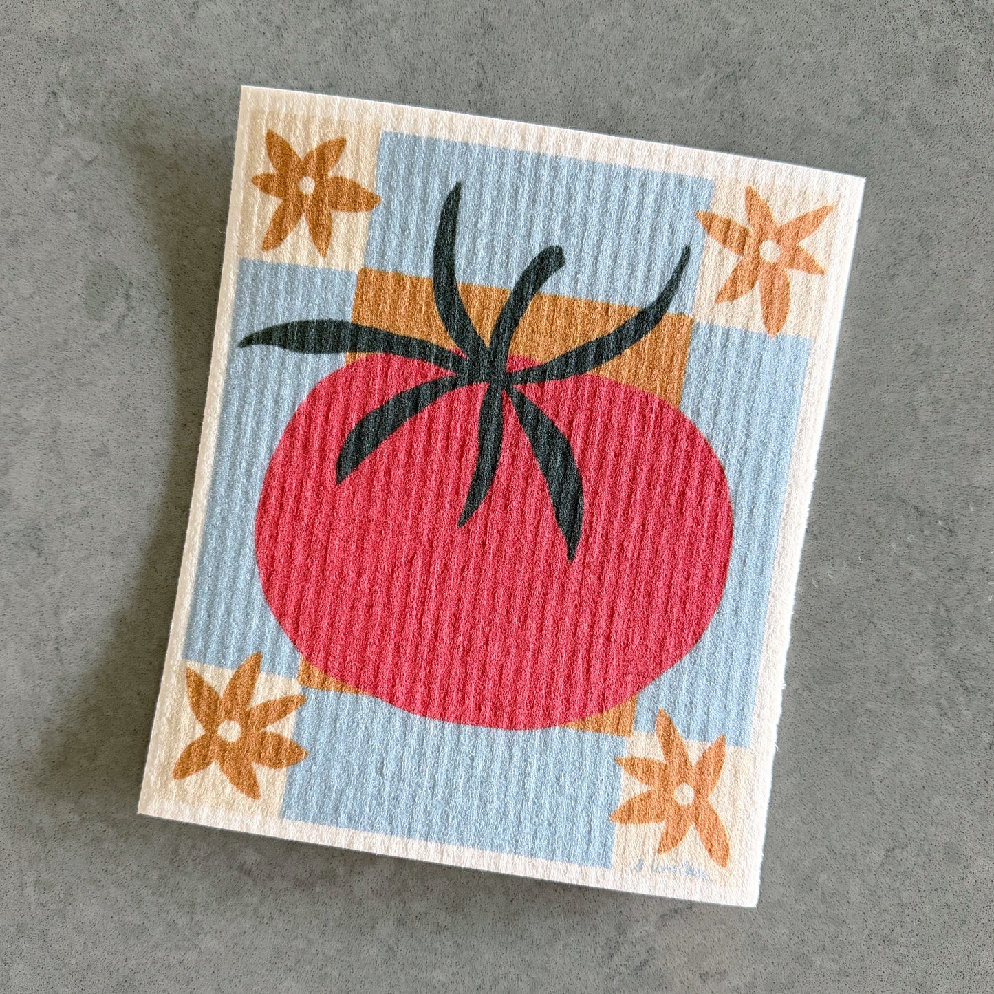A square textured paper dishcloth featuring a retro-style illustration of a red tomato with dark green stem details on a light blue background, decorated with orange starburst flowers in the corners, shown on a gray surface.