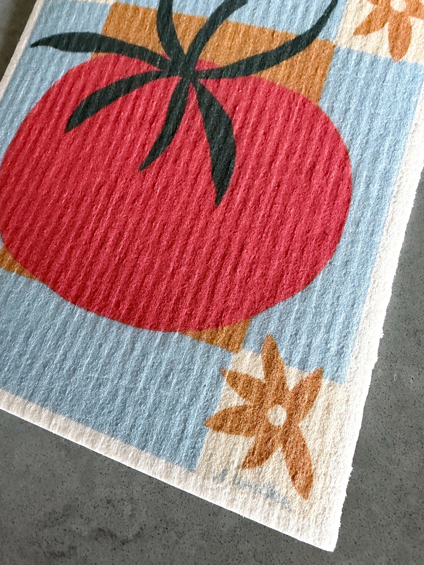 A close-up of a textured Swedish dishcloth featuring a large red tomato with a dark green stem on a light blue background, accented with orange flowers and geometric shapes.