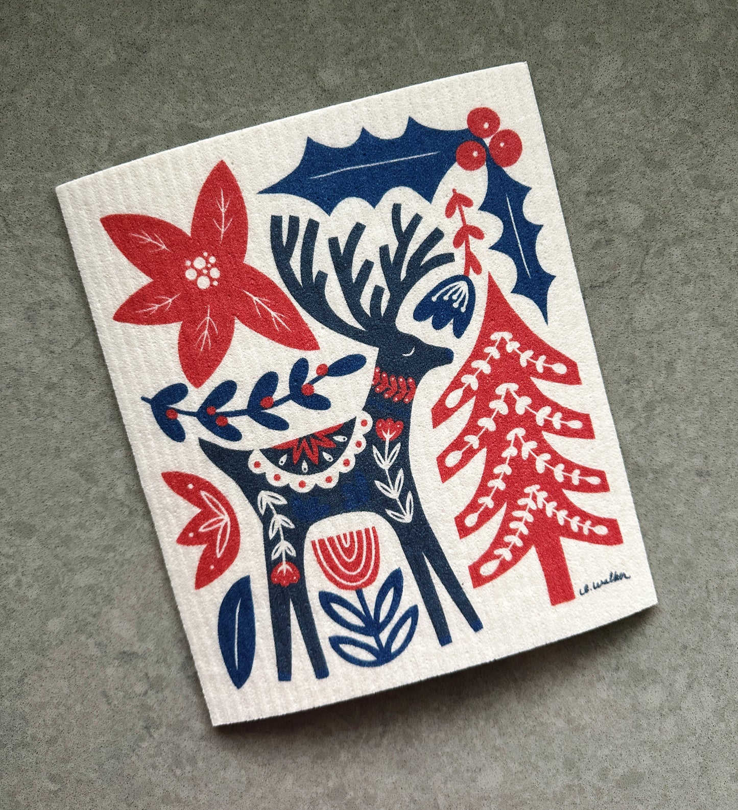 Decorative dishcloth with a festive design featuring a deer, holly, and Christmas tree on a gray background.