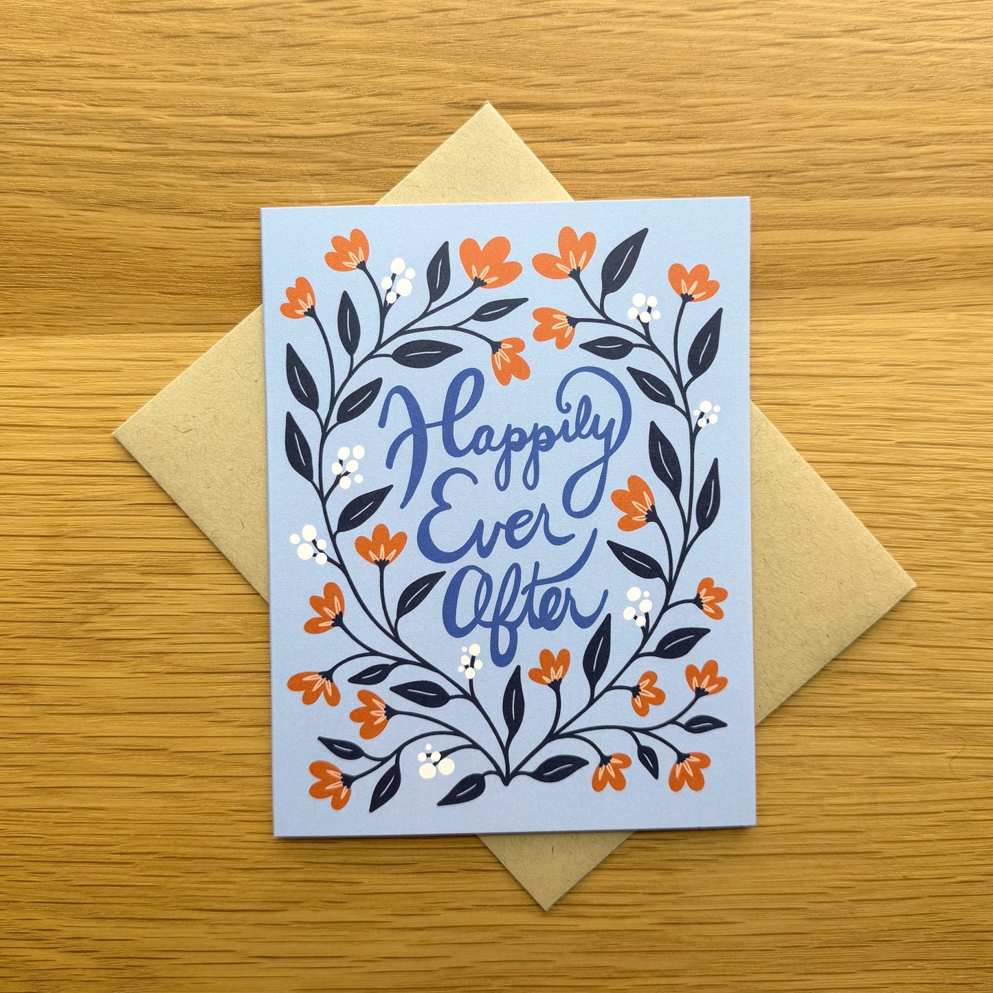 A greeting card with a light blue background and a floral wreath design in orange, navy, and white, with the words 'Happily Ever After' in blue script. The card is placed on top of a kraft paper envelope on a light wood surface.