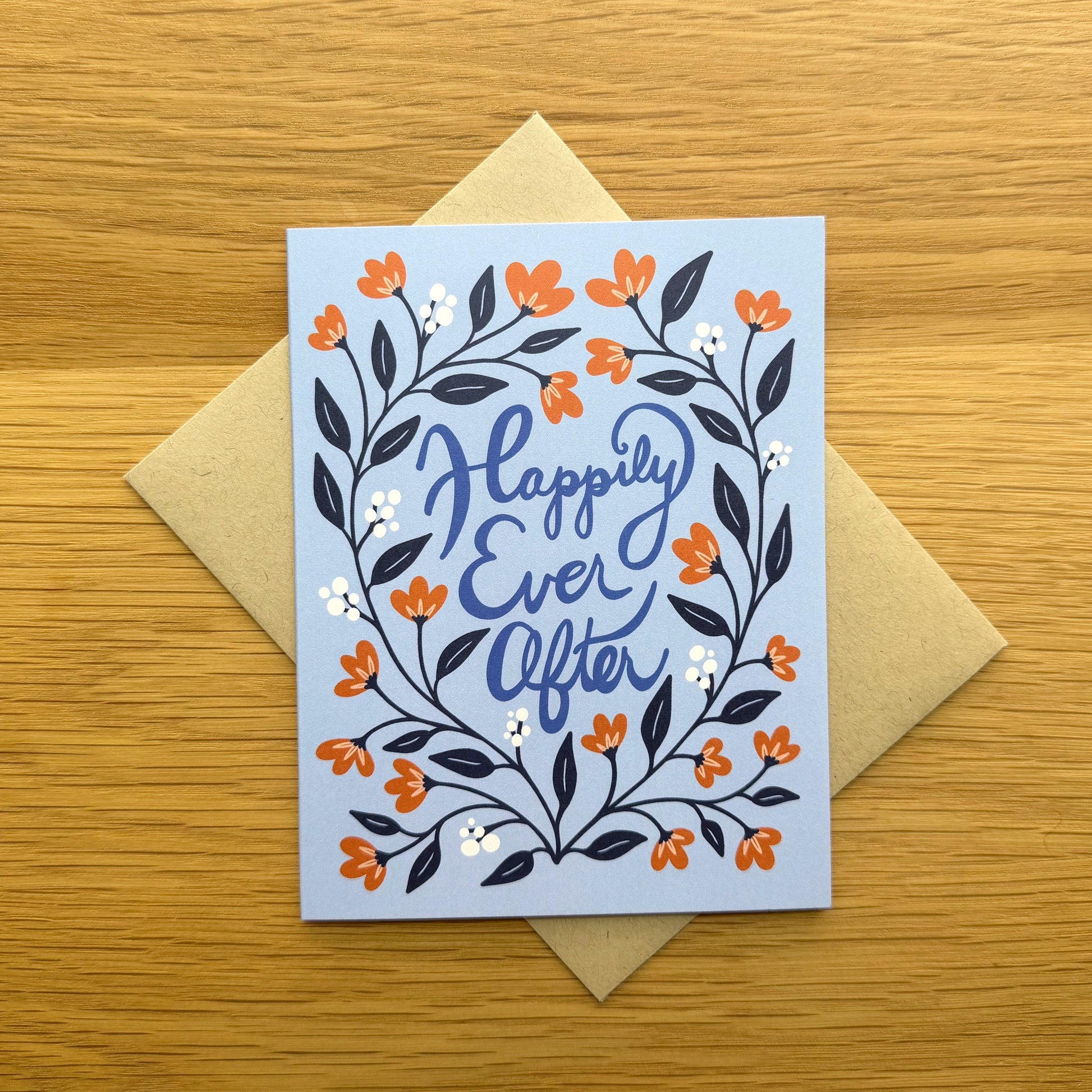 A greeting card with a light blue background and a floral wreath design in orange, navy, and white, with the words 'Happily Ever After' in blue script. The card is placed on top of a kraft paper envelope on a light wood surface.